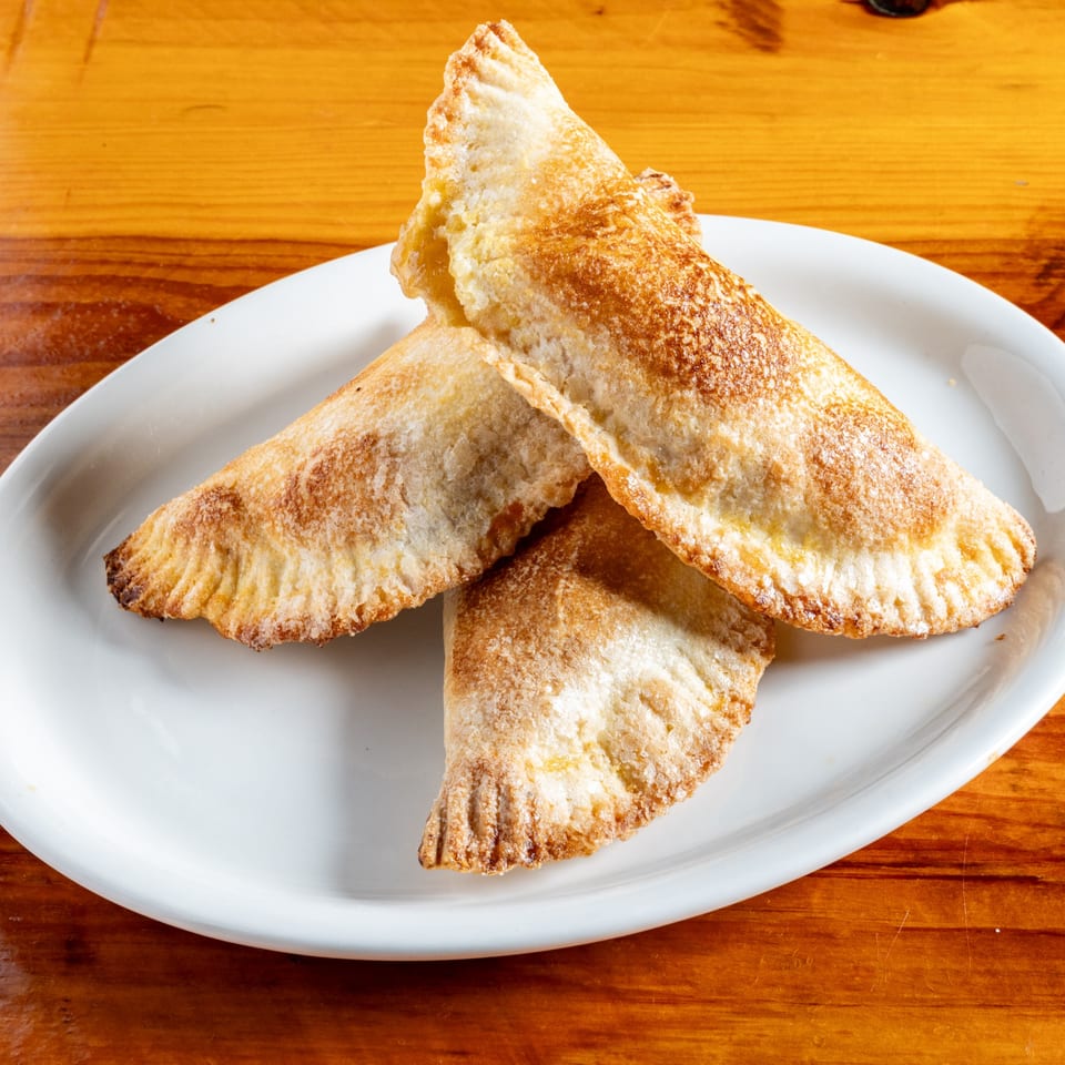 Three golden-brown Mexican empanadas with crimped edges on a white plate, on a wooden table.