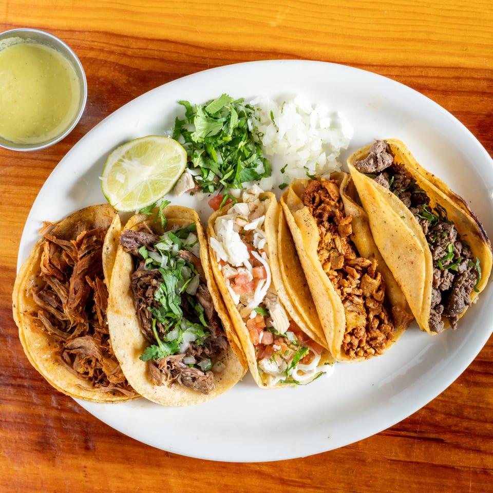A white plate of five diverse El Rey Del Grill tacos, lime, cilantro, and onions on a wooden table.