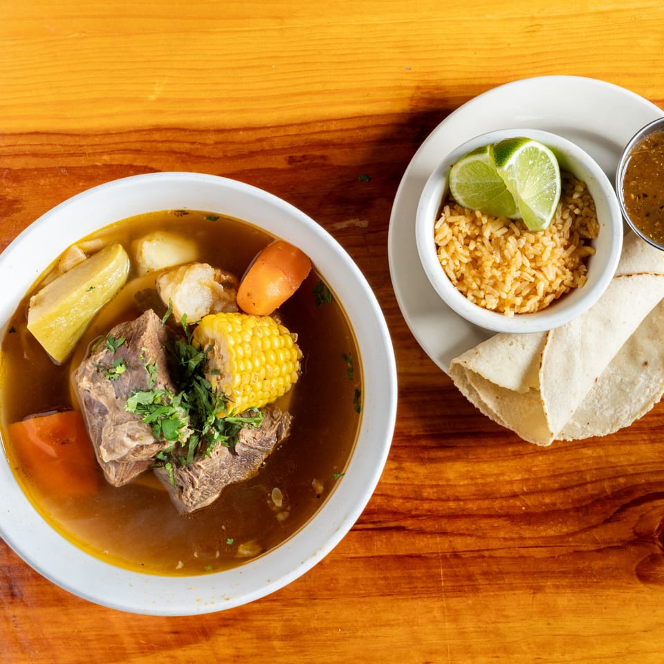 Hearty Mexican beef soup with corn, carrots, and potatoes, served with rice, lime, and tortillas.